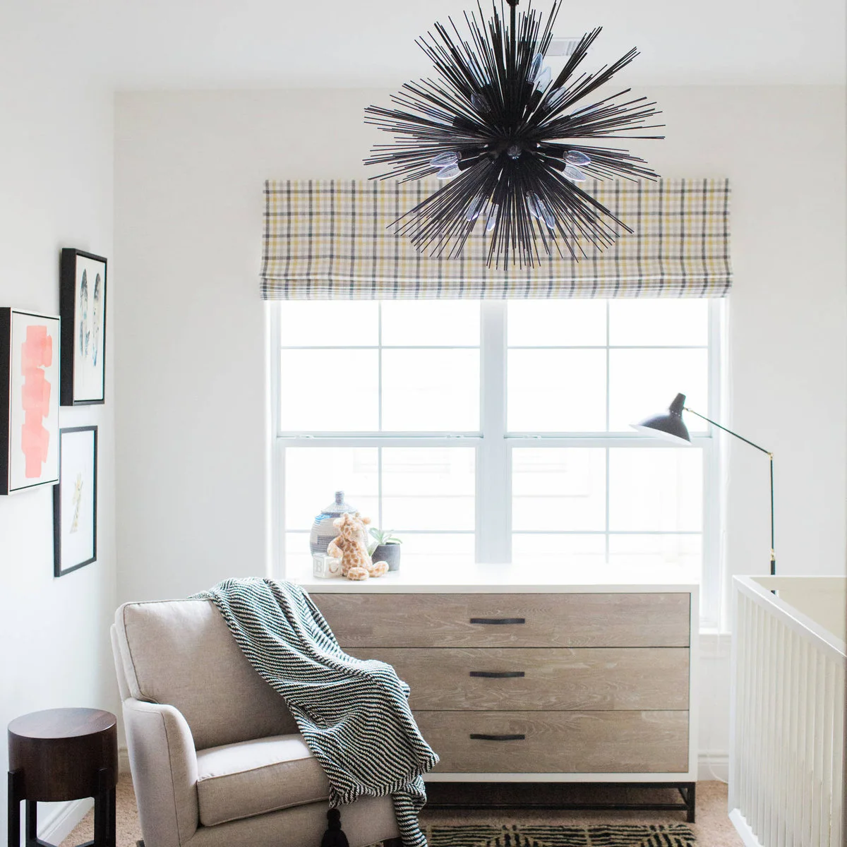 Neutral nursery corner with a pale wood dresser, upholstered chair, plaid blind and dark sculptural ceiling light