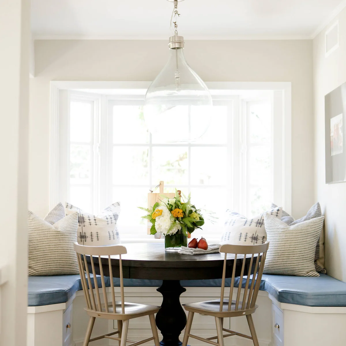A blue-cushioned wraparound banquette with striped pillows, a dark pedestal table and two pale wooden chairs.