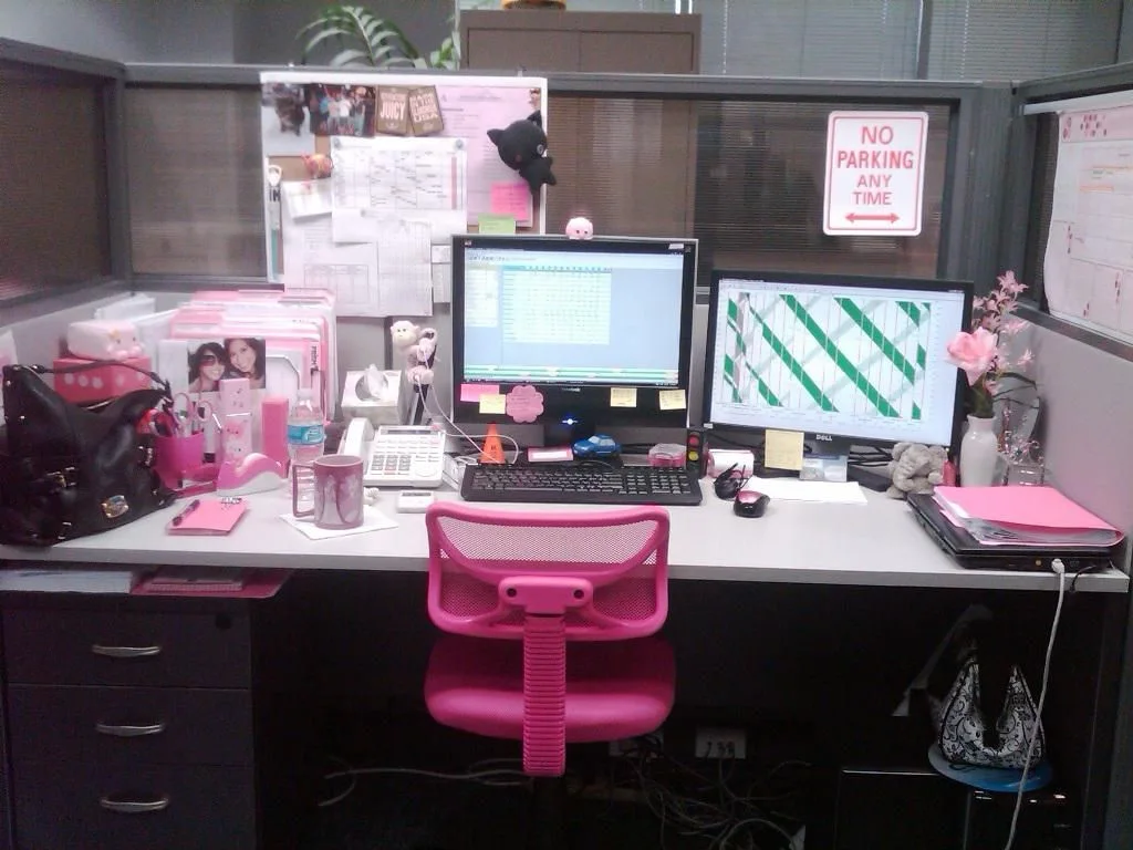 Pink office chair, files and desk accessories in a gray cubicle with two monitors and a notice board.