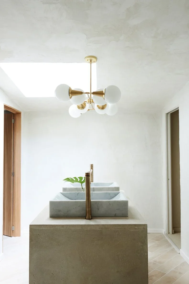 Two pale rectangular vessel basins stand along a broad gray block-like washstand beneath a skylight and a gold-toned globe light beside plain pale walls