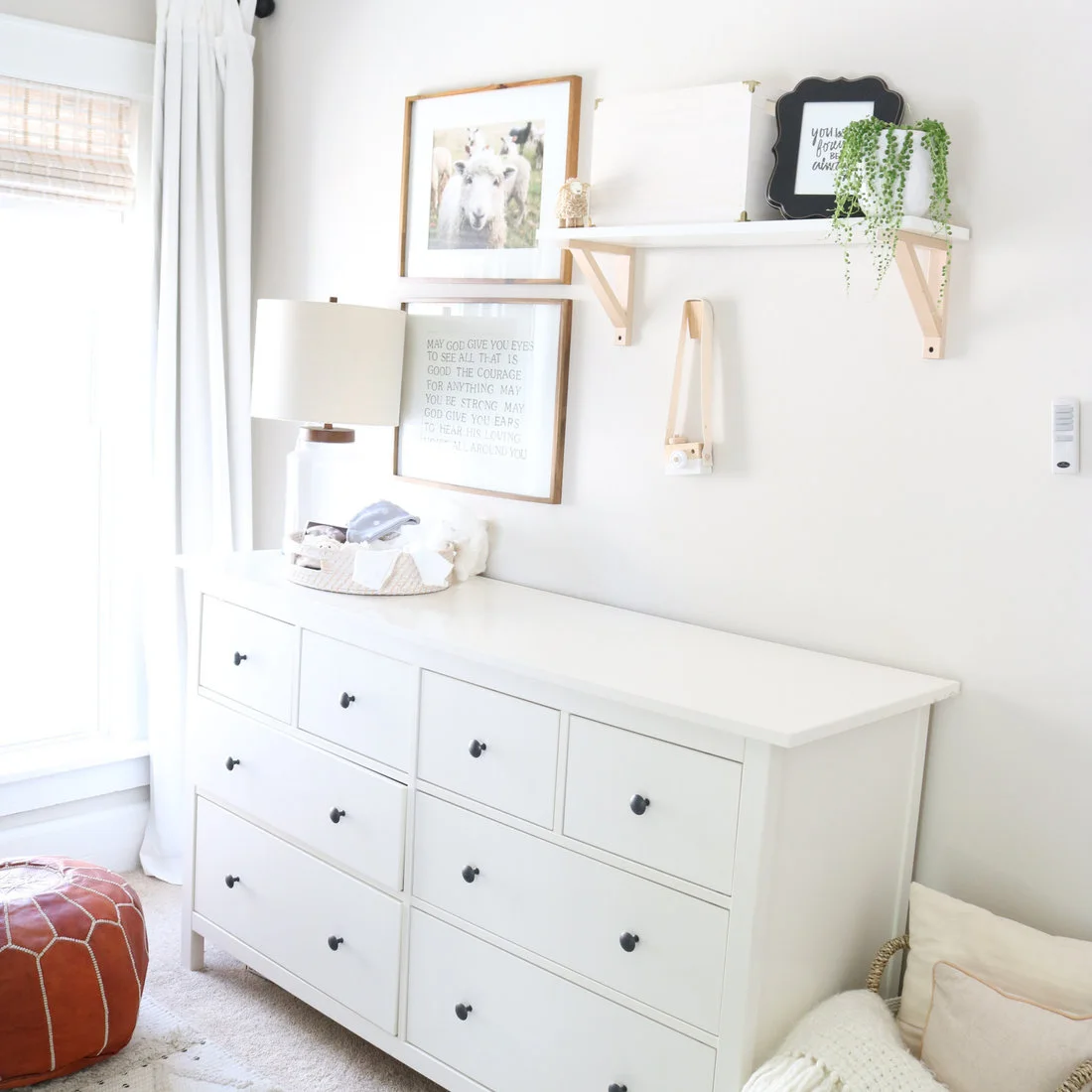 A broad white dresser with black knobs sits below a wood shelf beside pale curtains, a small lamp, framed pictures and a plant.