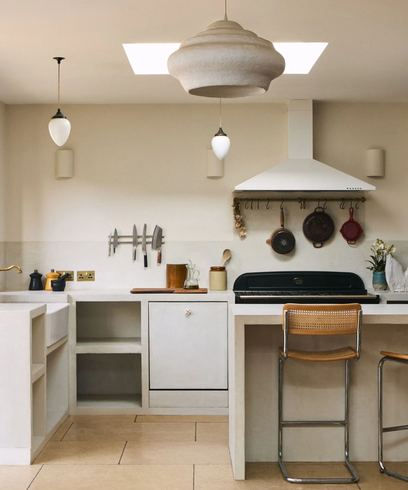 A pale kitchen has open lower compartments, a black cooker, hanging pans, a knife rail and woven-back stools beneath pale pendant lights
