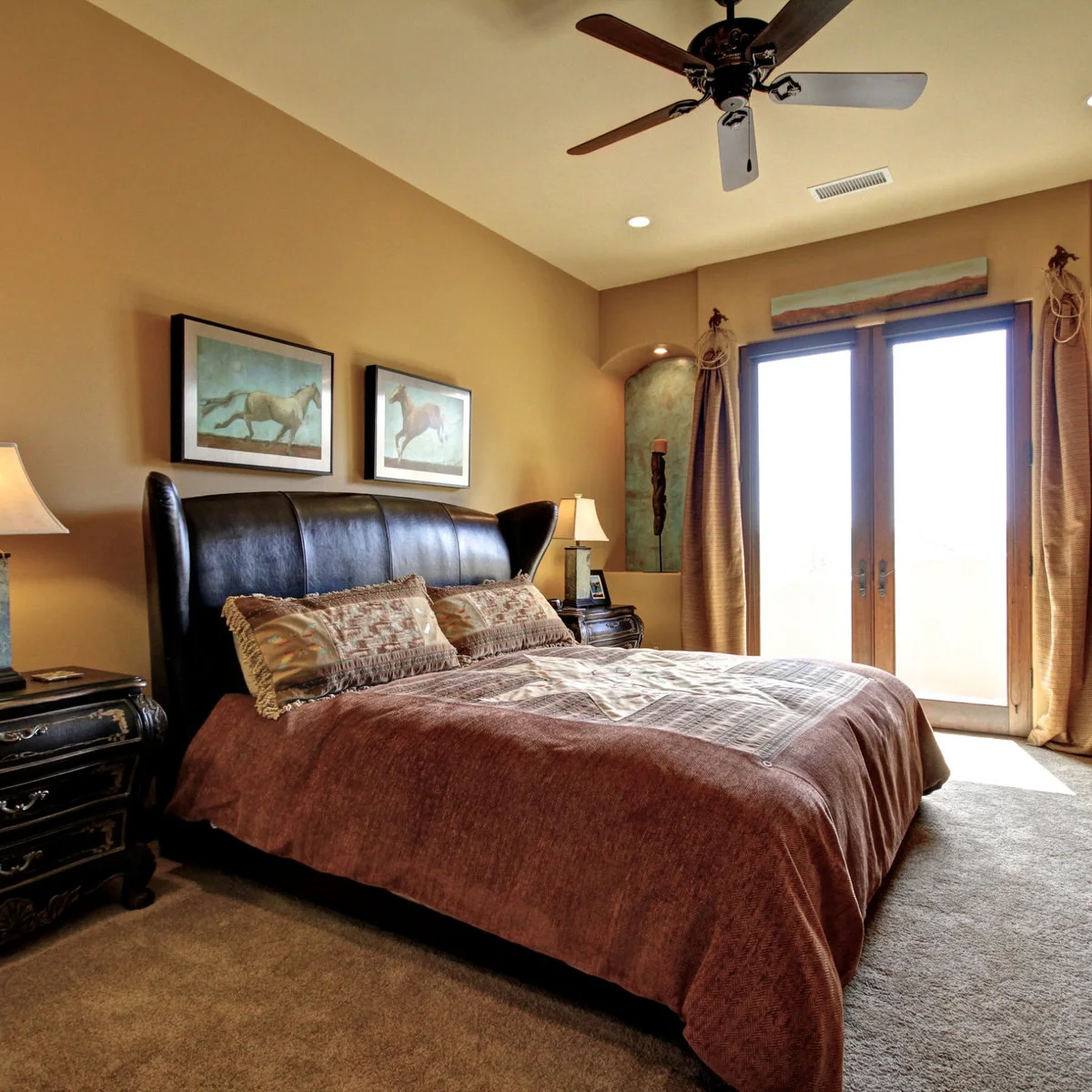 Western bedroom with a dark winged headboard, warm tan walls, two horse prints and long curtains by glazed doors