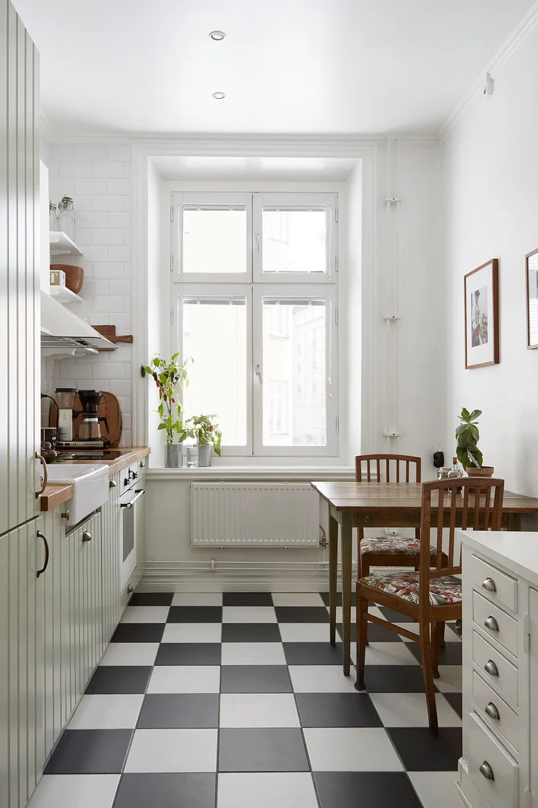 A narrow pale kitchen has a wood table and chairs, gray-and-white checkerboard floor, a tall bright window and a simple working run on the opposite wall.
