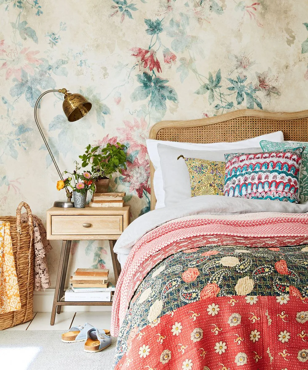 Soft pink and blue floral wallpaper behind a cane headboard with colorful pillows and layered quilts.