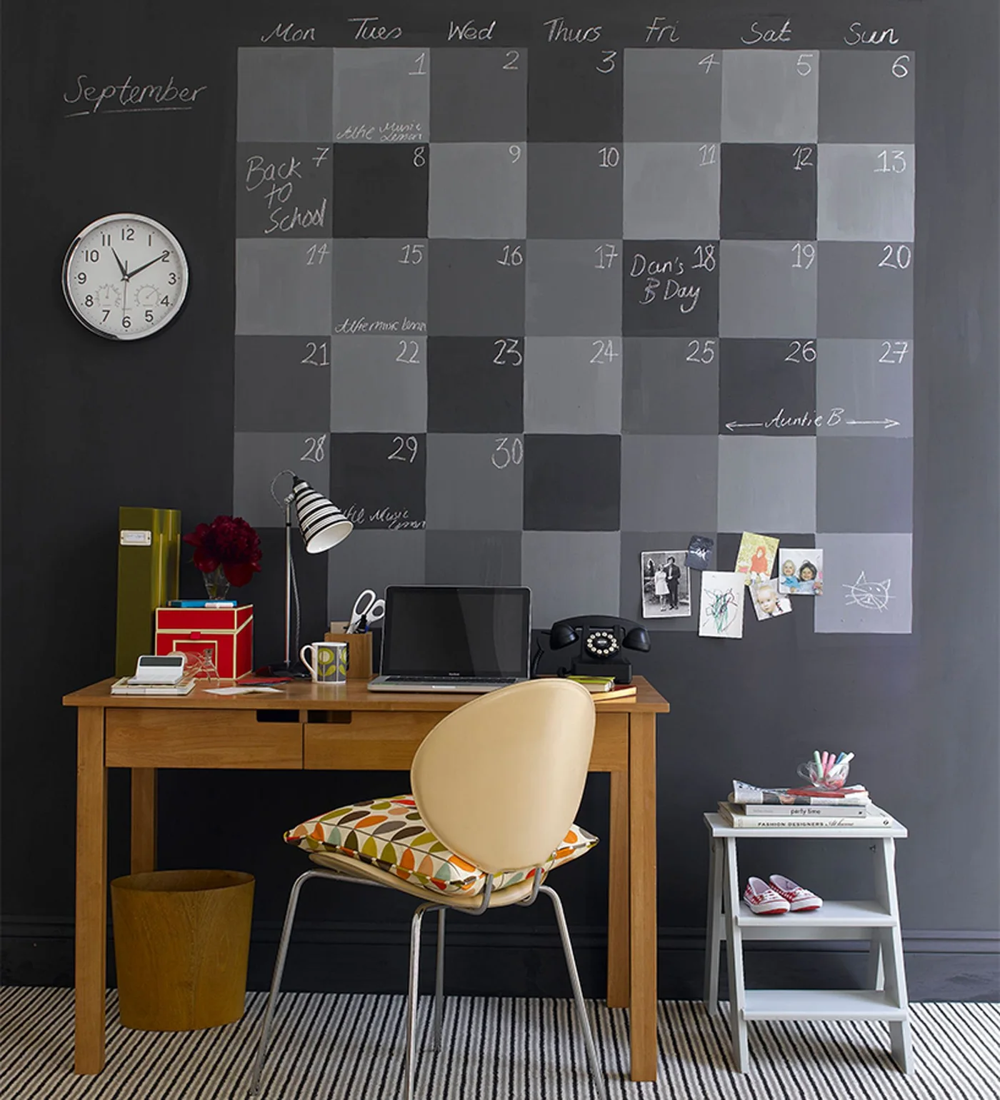A plain wood desk and pale chair stand beneath a large gray chalkboard calendar, with a striped lamp, a round clock and small colorful storage boxes.