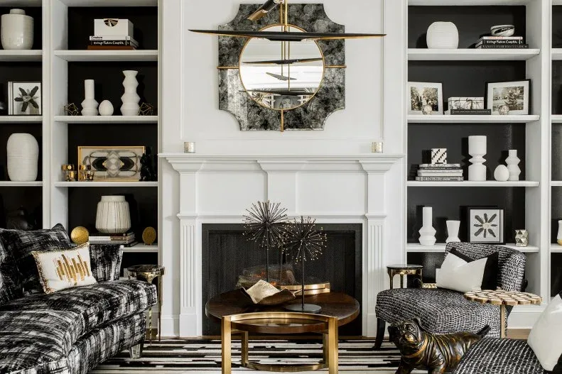 Living room with white fireplace, dark-backed shelves, white decorative objects, round gold-framed mirror, black-and-white patterned chairs and round golden table.
