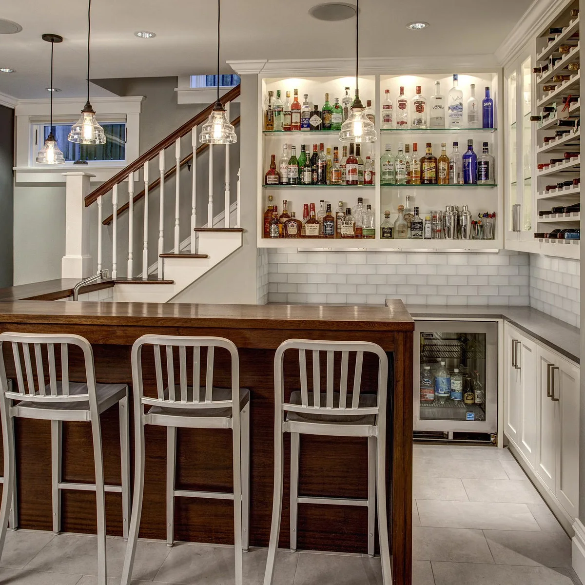 White basement bar with wood-colored raised counter, three stools and illuminated bottle shelves