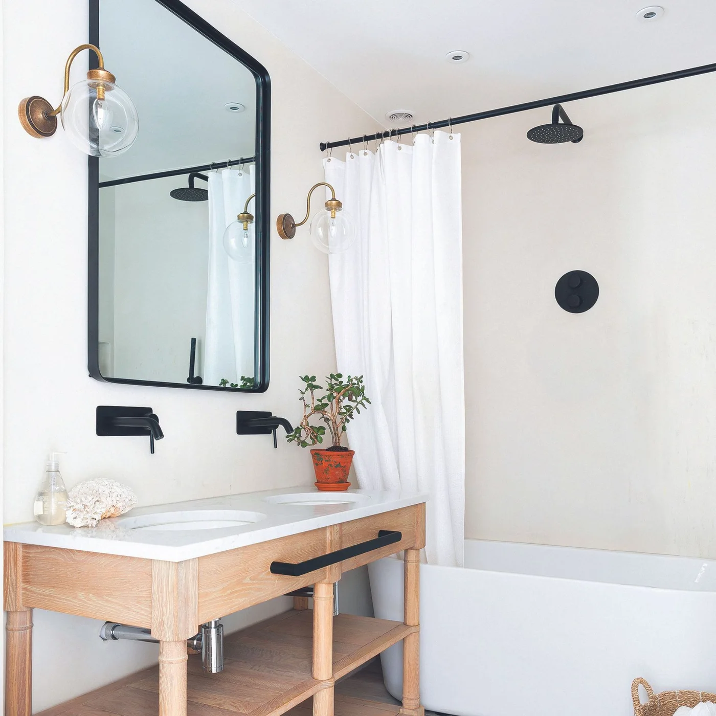 Two white basins sit in a wood console below a wide black-framed mirror, beside a bath with a white shower curtain and black shower fittings.