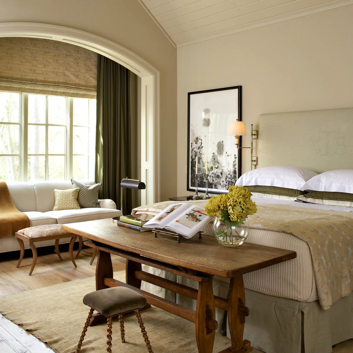 Pale bedroom with a settee beneath an arched window and a work table near the bed