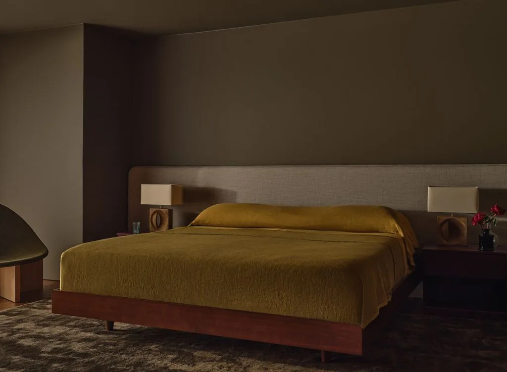Earthy olive bedroom walls with a wide gray headboard, low wooden bed, mustard-toned bedding, matching bedside lamps, and dark patterned rug.