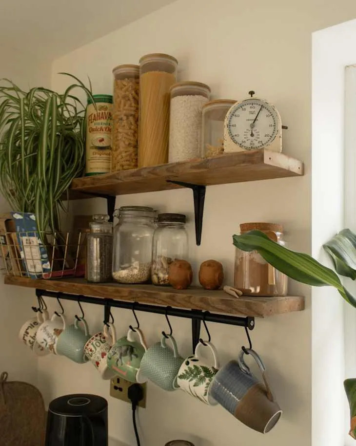 Two rustic wooden kitchen shelves with jars, patterned mugs, a vintage timer and greenery