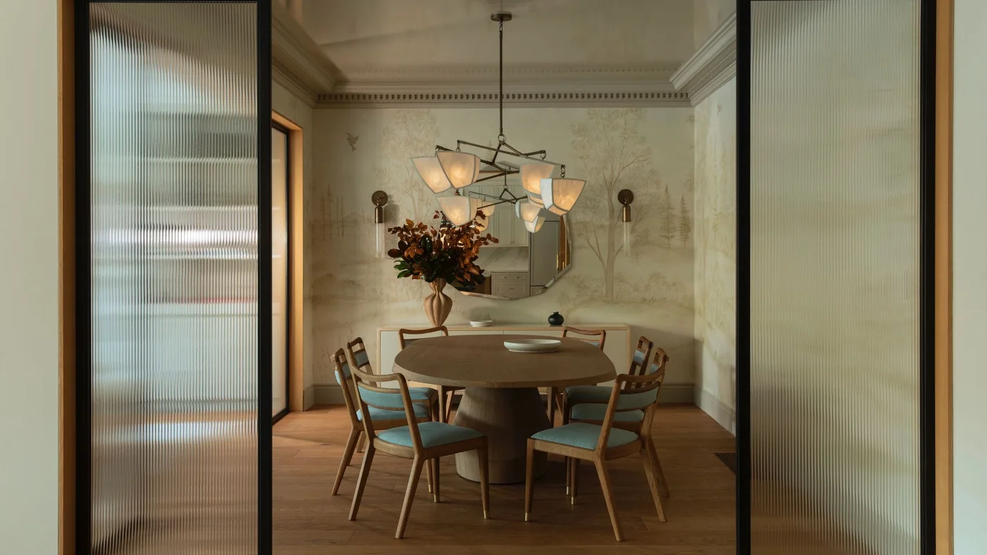 A pale sideboard behind a round dining table carries a tall dark-leaf arrangement toward one end and smaller vessels beneath a round mirror.