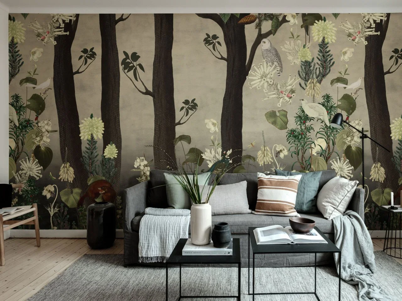 A gray sofa and slim dark tables sit in front of a large botanical wall mural with trees, flowers and birds.