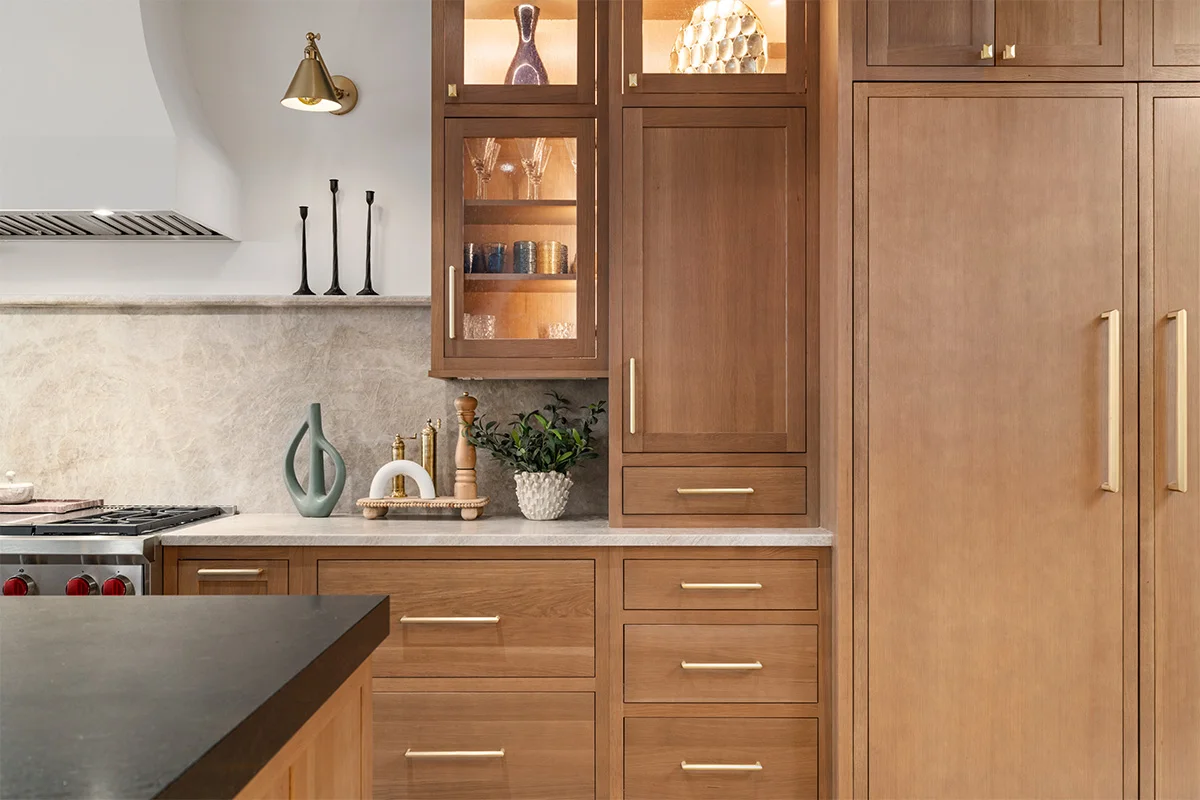 A glass-front display cupboard with warm lighting sits beside stained white oak drawers, a countertop cabinet and a tall paneled appliance.