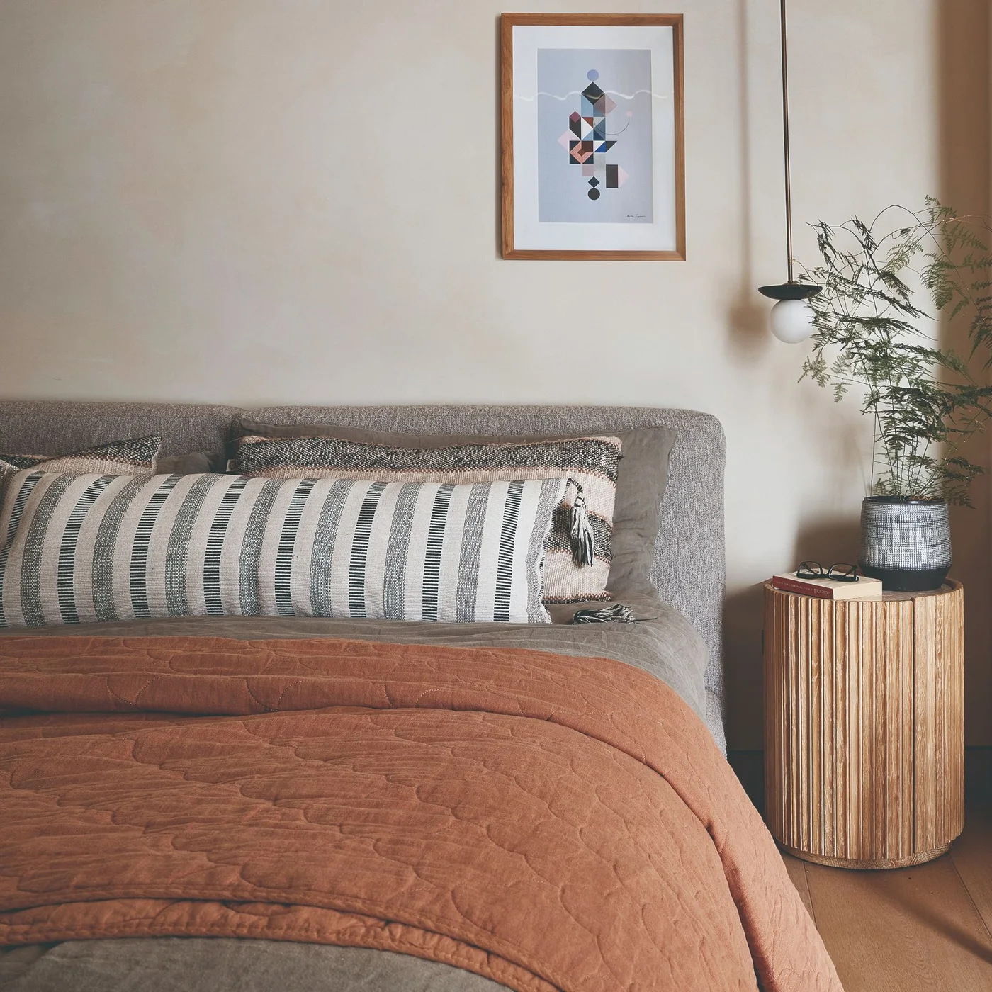 A gray upholstered bed has a long striped cushion and a rust-colored quilt beside a ribbed wood side table and a hanging globe light.