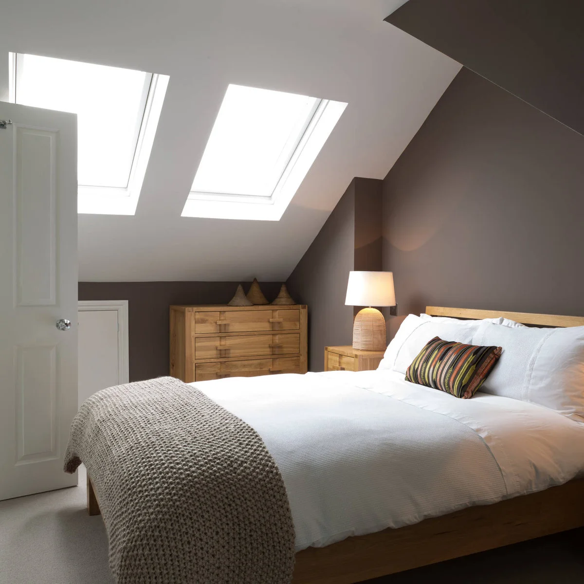 Brown attic bedroom walls below white roof slopes with wooden furniture and white bedding.