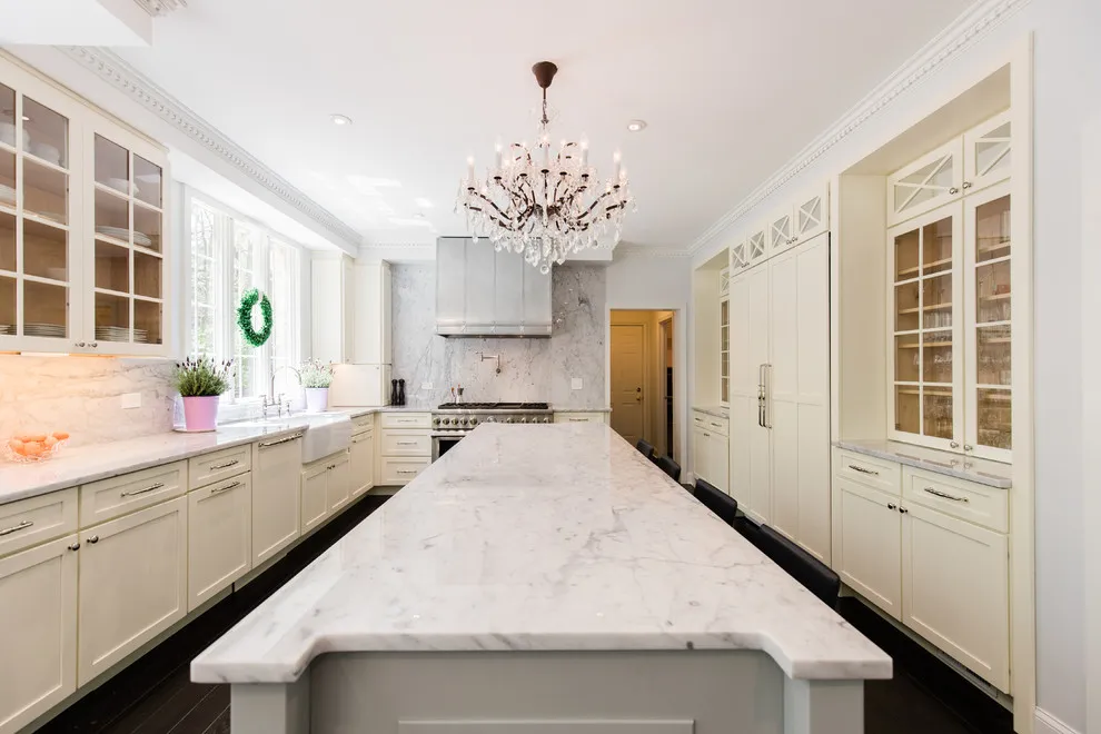 Pale framed cabinets with glass upper doors surround a long white island beneath a decorative chandelier and metal hood.
