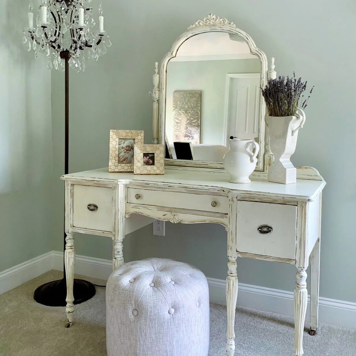 A distressed pale dressing table and ornate mirror stand above a round upholstered stool
