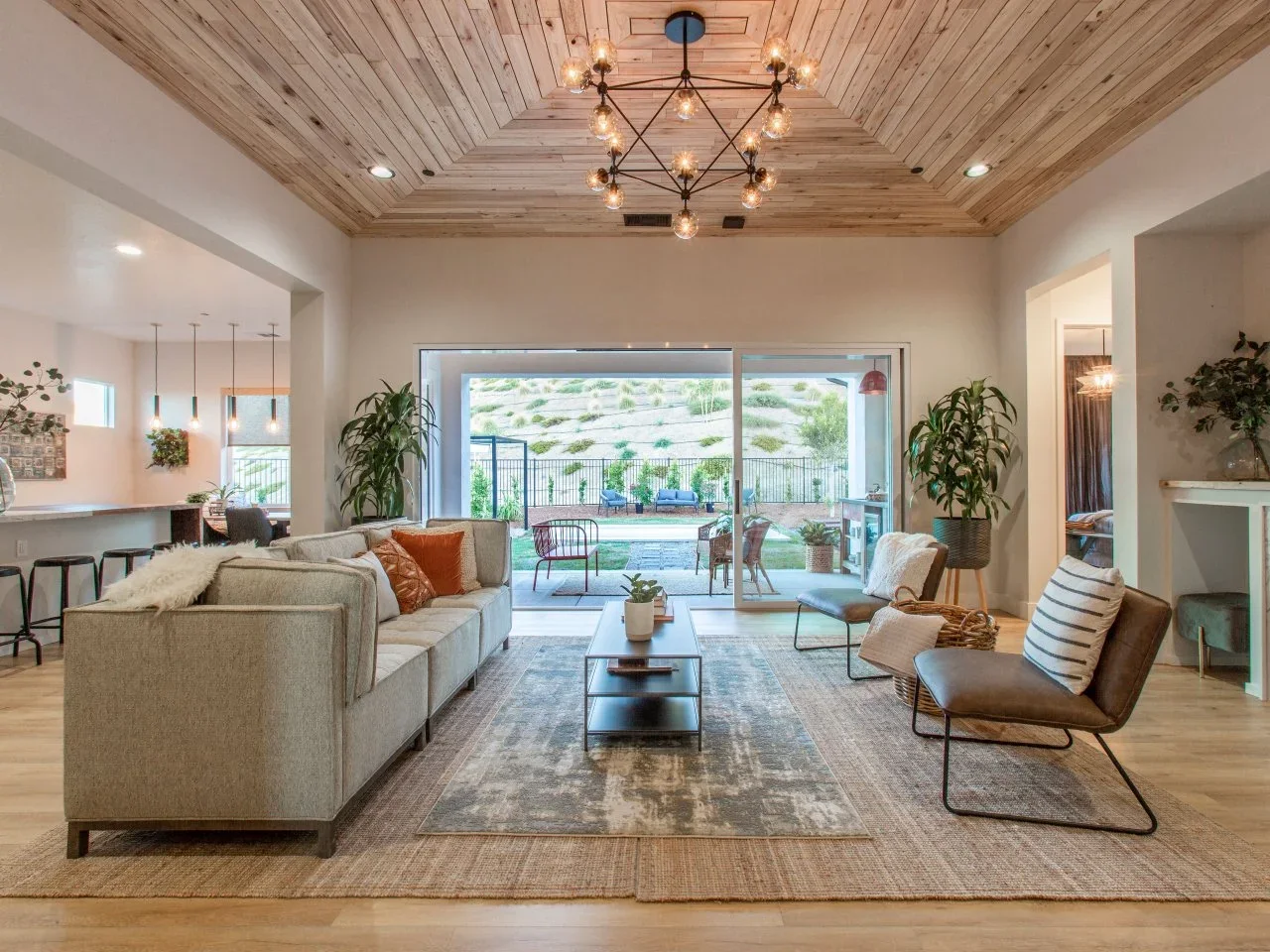 A gray sofa and brown chairs sit on layered rugs beneath a timber-clad ceiling beside garden doors.