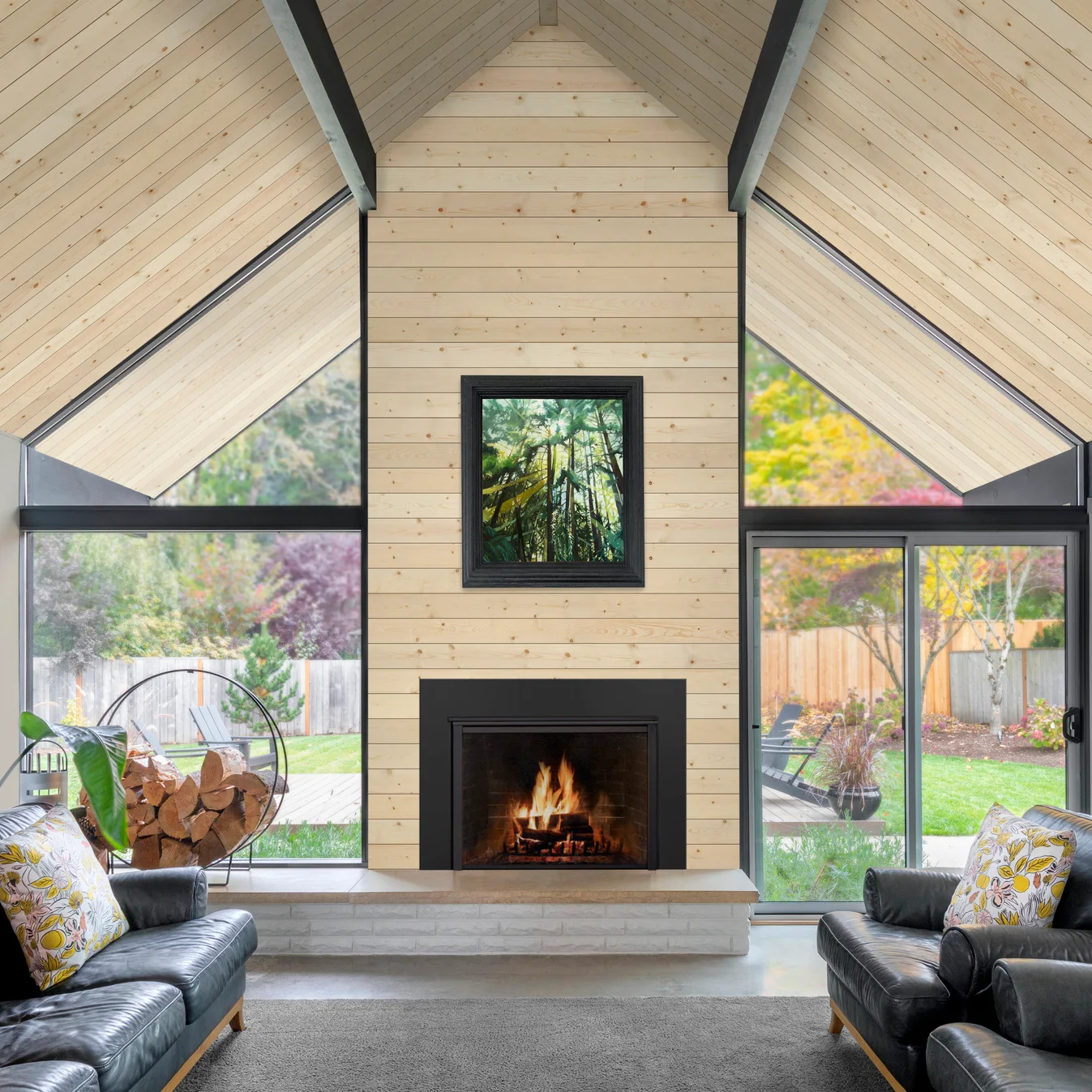 A pale wood-board fireplace rises between tall black-framed windows beneath a matching pitched ceiling, with dark seating and a raised pale hearth.