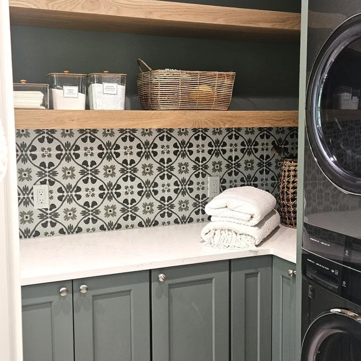Gray cabinets and a white counter sit beneath wood shelves and a black-and-white backsplash with green wall color beside stacked dark machines.