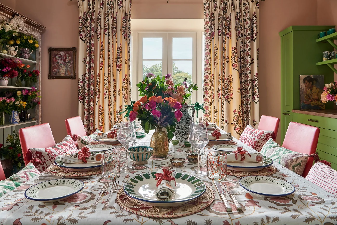 Floral curtains and a patterned tablecloth surround pink dining chairs beside green storage and a display of flowers