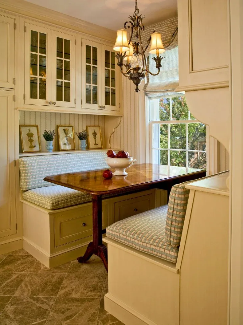 Two pale patterned booth benches with drawer fronts beneath glass-front display cabinets, a wooden pedestal table, shaded chandelier, framed pictures, and a tall window.