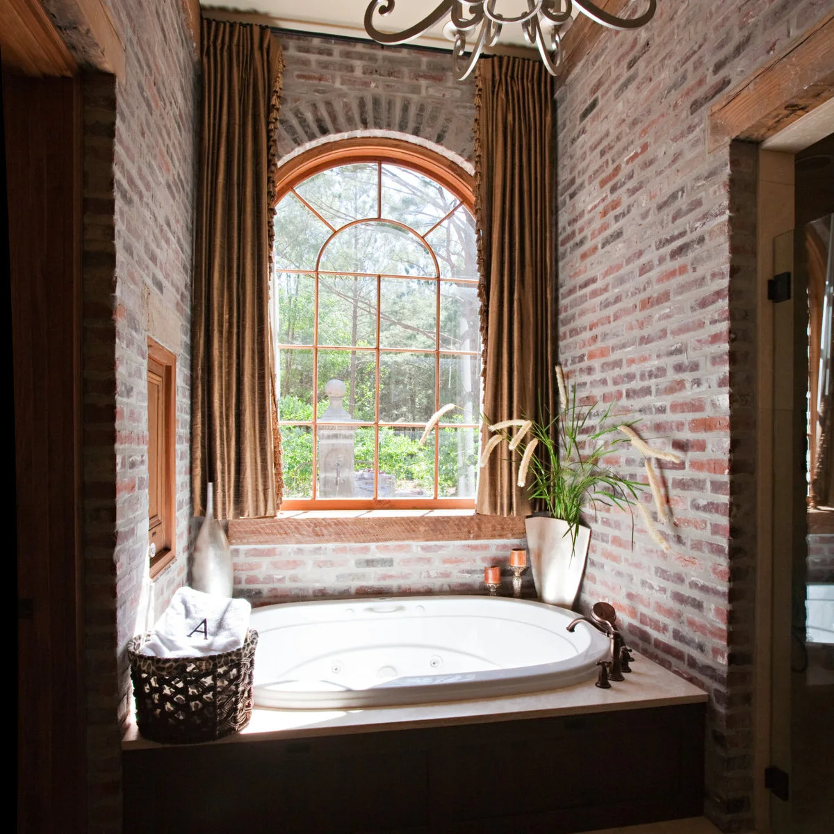 Brown full-length curtains frame an arched window above a built-in bath in a brick-walled bathroom.