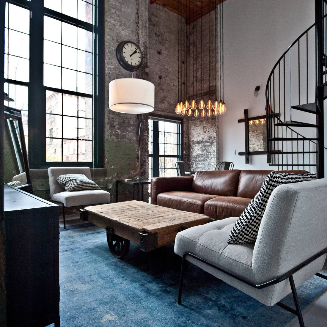 Two pale chairs and a brown sofa around a wheeled wood table beside tall factory windows