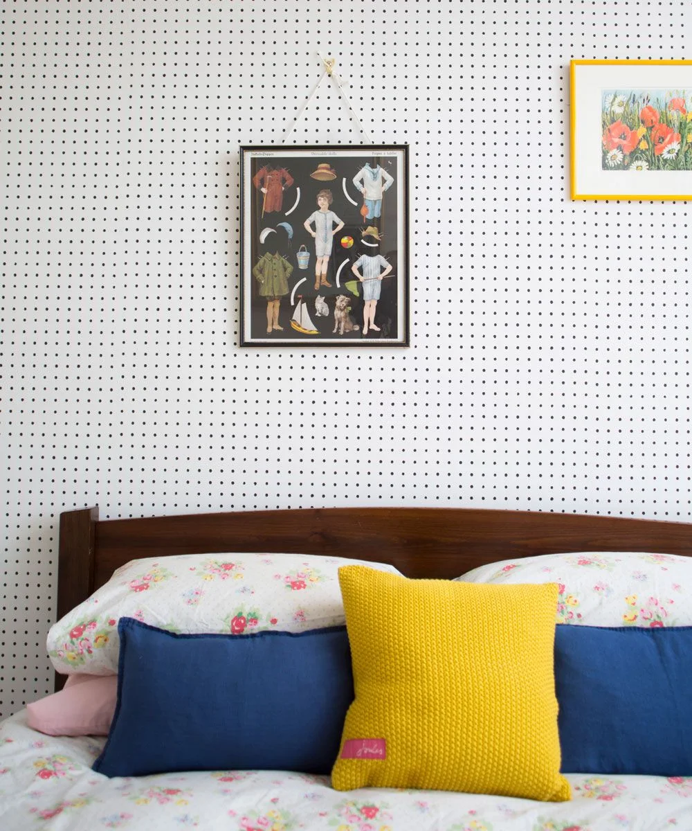 Tiny black dots cover a white wall above a dark wood bed with floral bedding, navy cushions, a yellow knitted cushion and two framed pictures.
