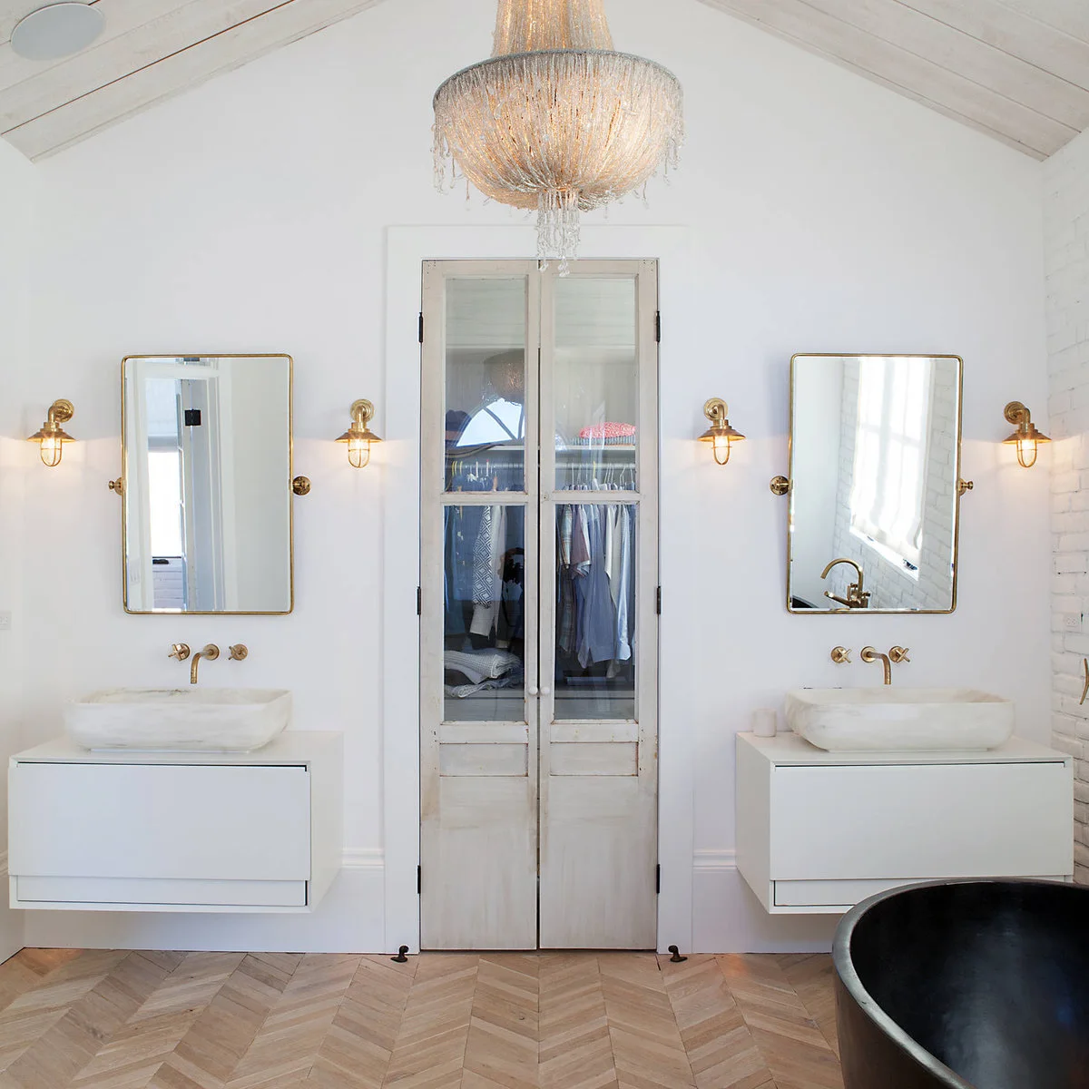 Weathered glass-front cabinet doors stand between two white floating vanities with gold-colored taps and framed mirrors.