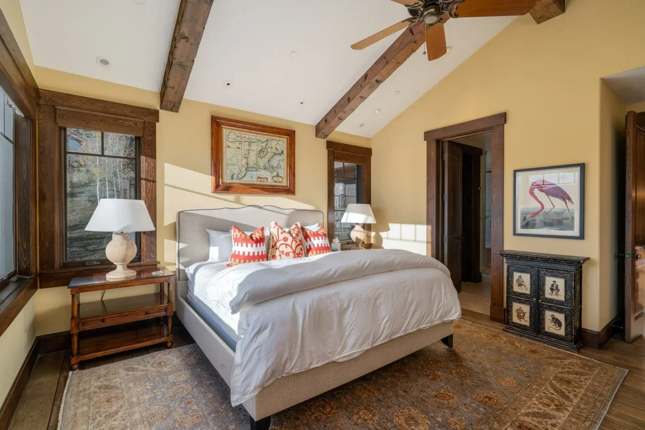 Warm yellow walls surround a pale upholstered bed beneath exposed wood beams, with rust-patterned pillows, wood tables and framed pictures.