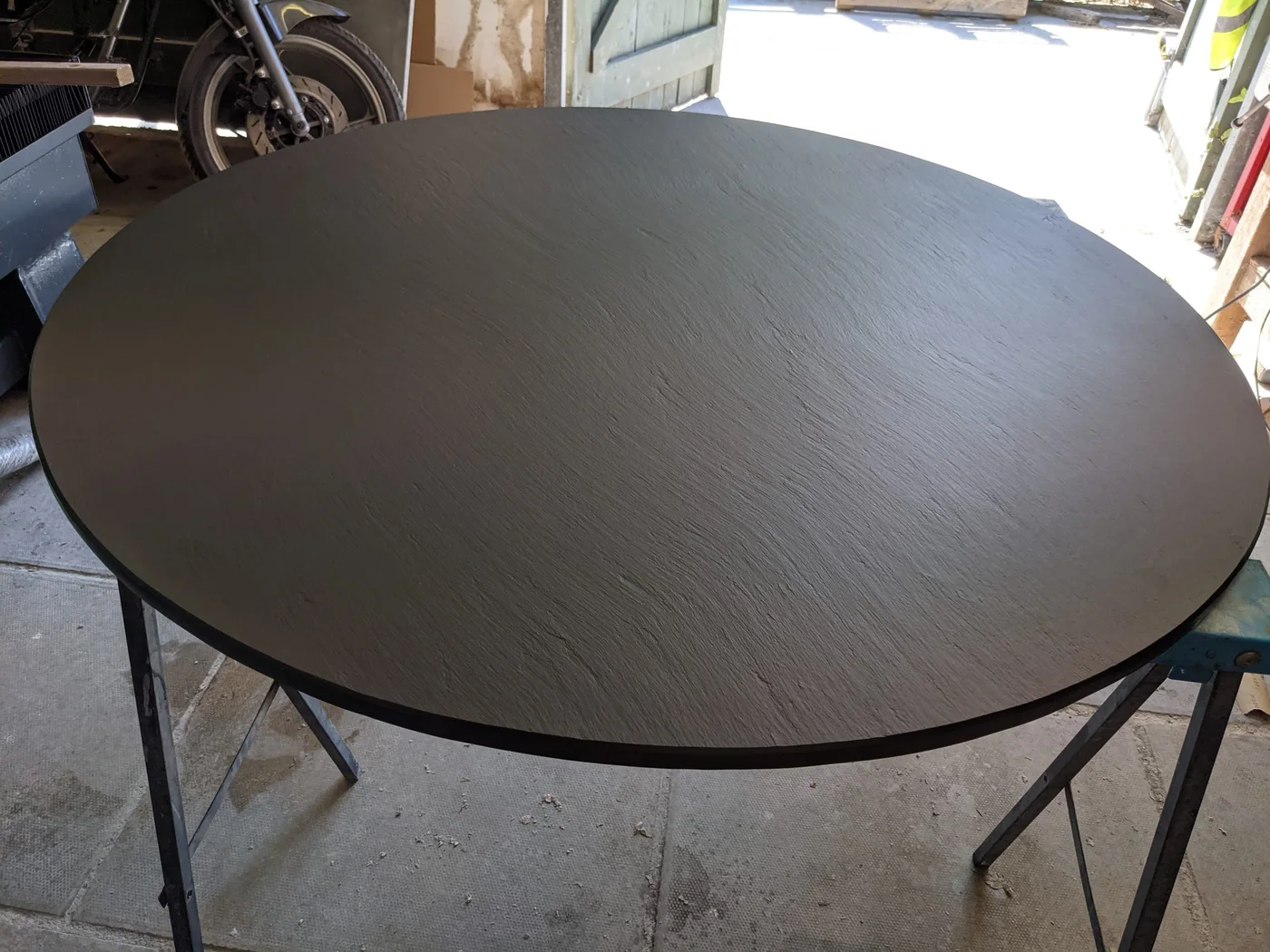A circular riven slate slab rests on workshop supports with its dark front edge visible.