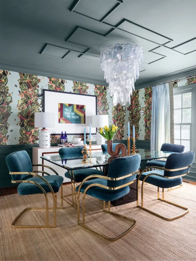 White dining sideboard with warm pink edging beneath artwork and white lamps behind teal chairs and a glass table
