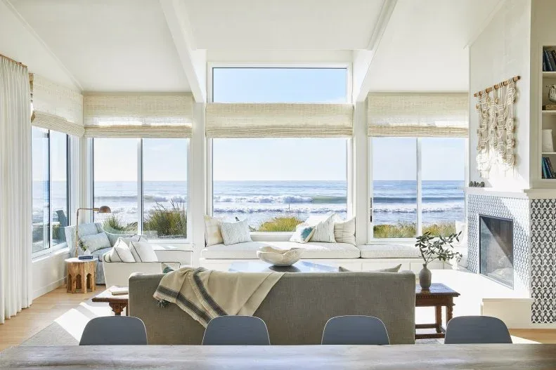Coastal room with large sea-view windows, low pale seating, woven-looking blinds and a patterned fireplace edge.
