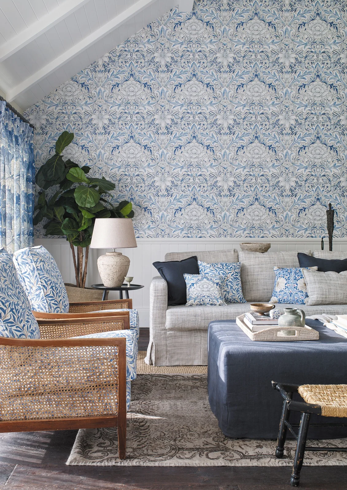 Blue-and-white scrolling wallpaper covers the wall above paneling behind a pale sofa under white roof beams.