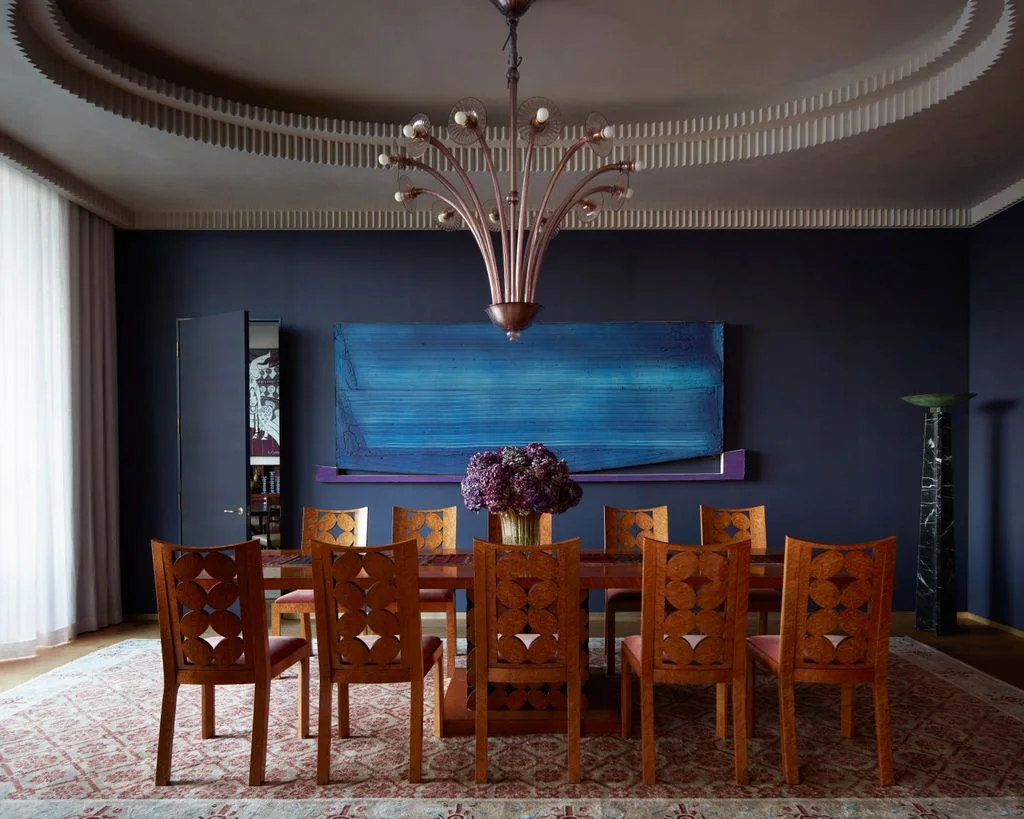 Carved wooden dining chairs surround a long table beneath a branching chandelier beside dark blue walls and large blue artwork