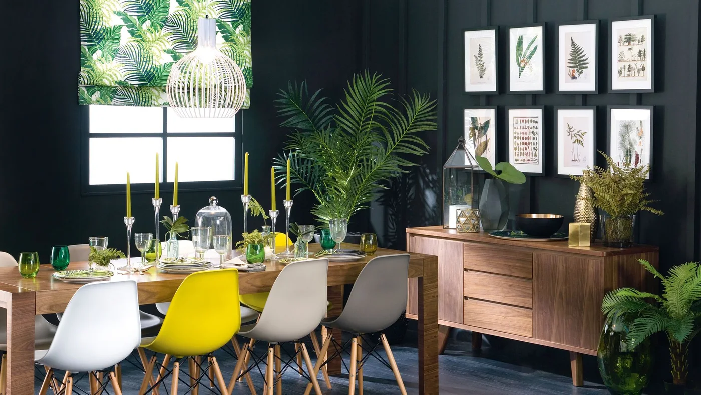 A wood sideboard holds a tall glass lantern, a faceted vase, a dark bowl and leafy stems beneath botanical prints on a dark wall.