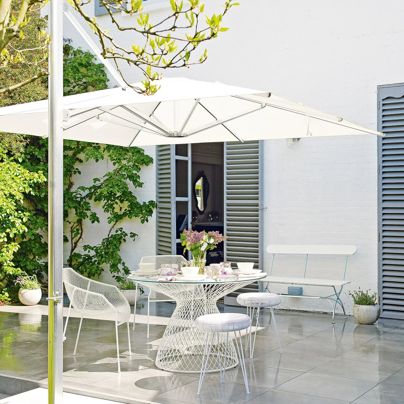 A pale umbrella shelters white outdoor dining furniture beside a white wall and gray shutters