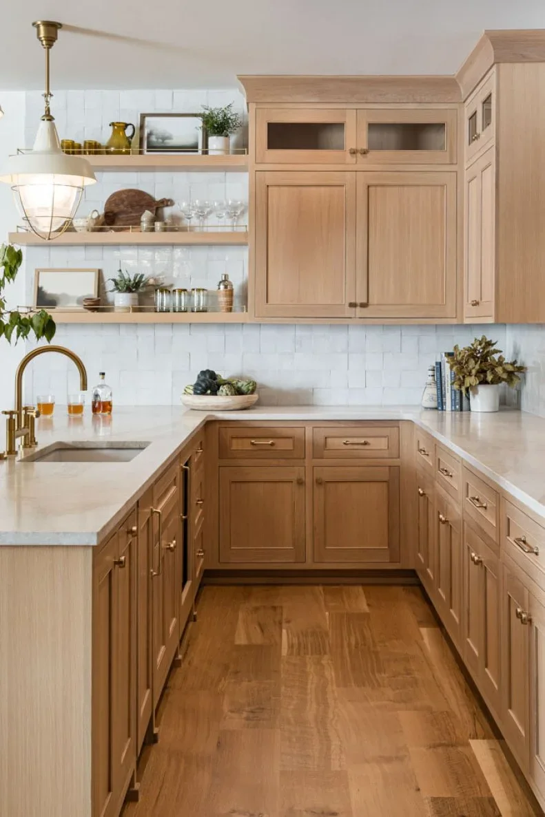 Light wood cabinets wrap a small kitchen corner beneath pale counters and white tile, with open shelves beside the sink.