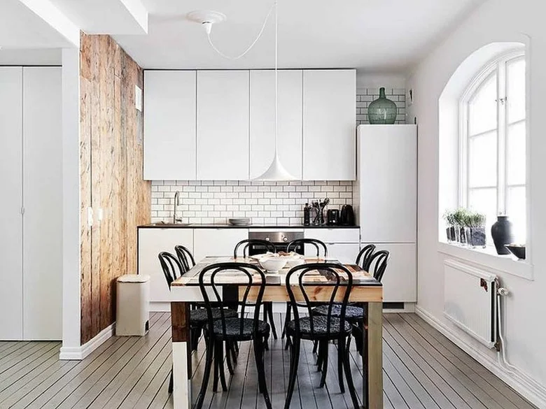 A compact white kitchen has flat upper cupboards, a tiled backsplash, a wood dining table with black bentwood chairs, a white pendant and pale floorboards.