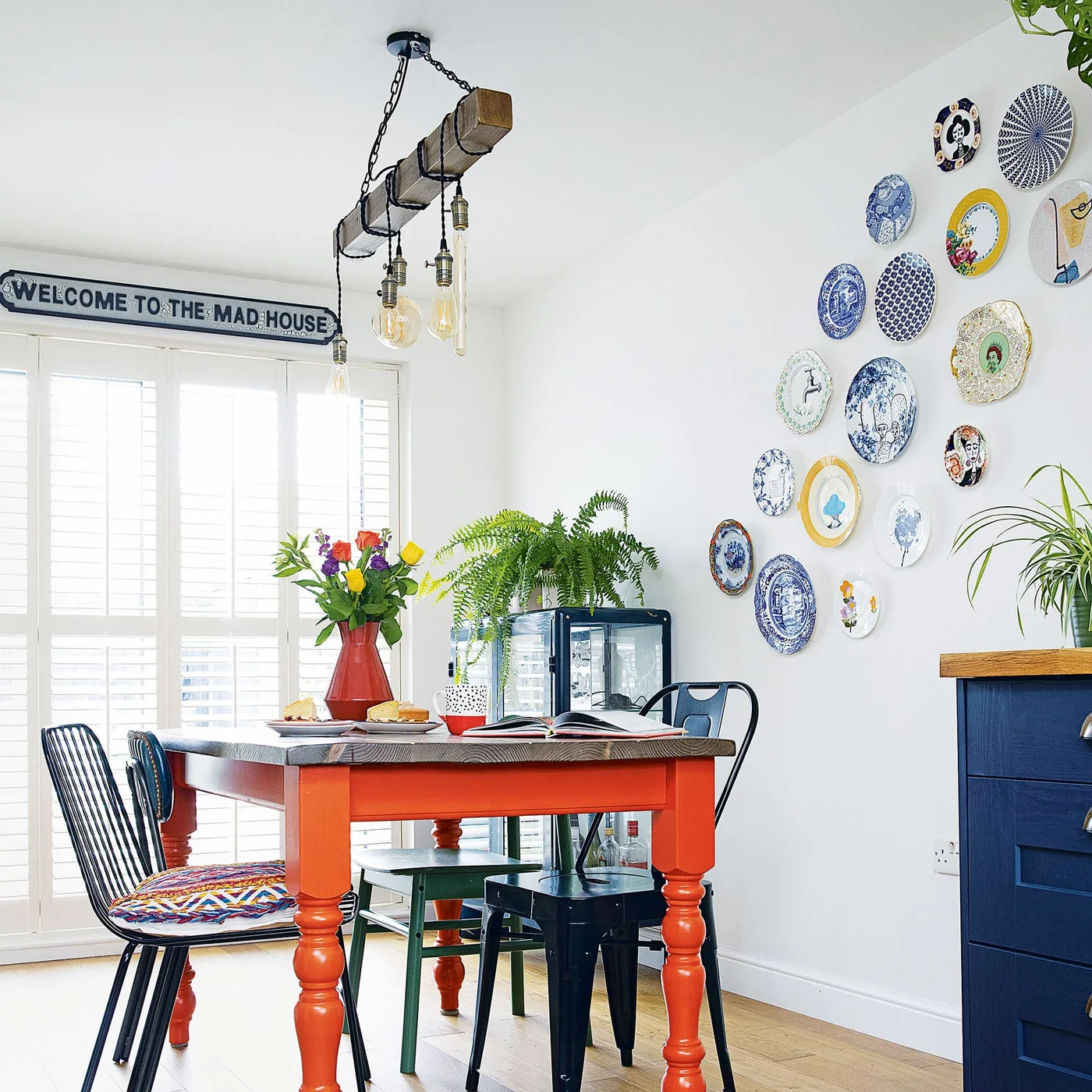 A small orange dining table and mixed dark chairs stand beneath a display of decorative plates beside blue cabinetry and shuttered doors.