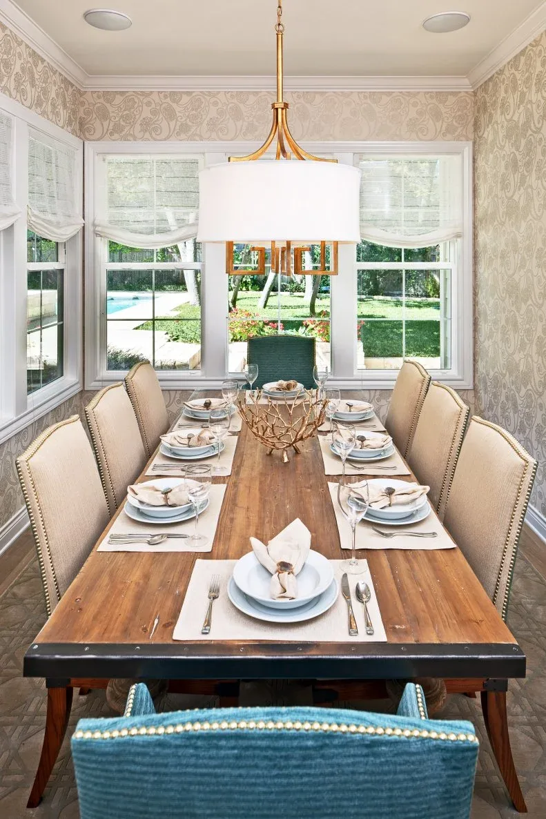 Pale upholstered chairs with nailhead edges beside a wooden dining table and a blue textured end chair, beneath a broad white-shaded light and surrounding windows.