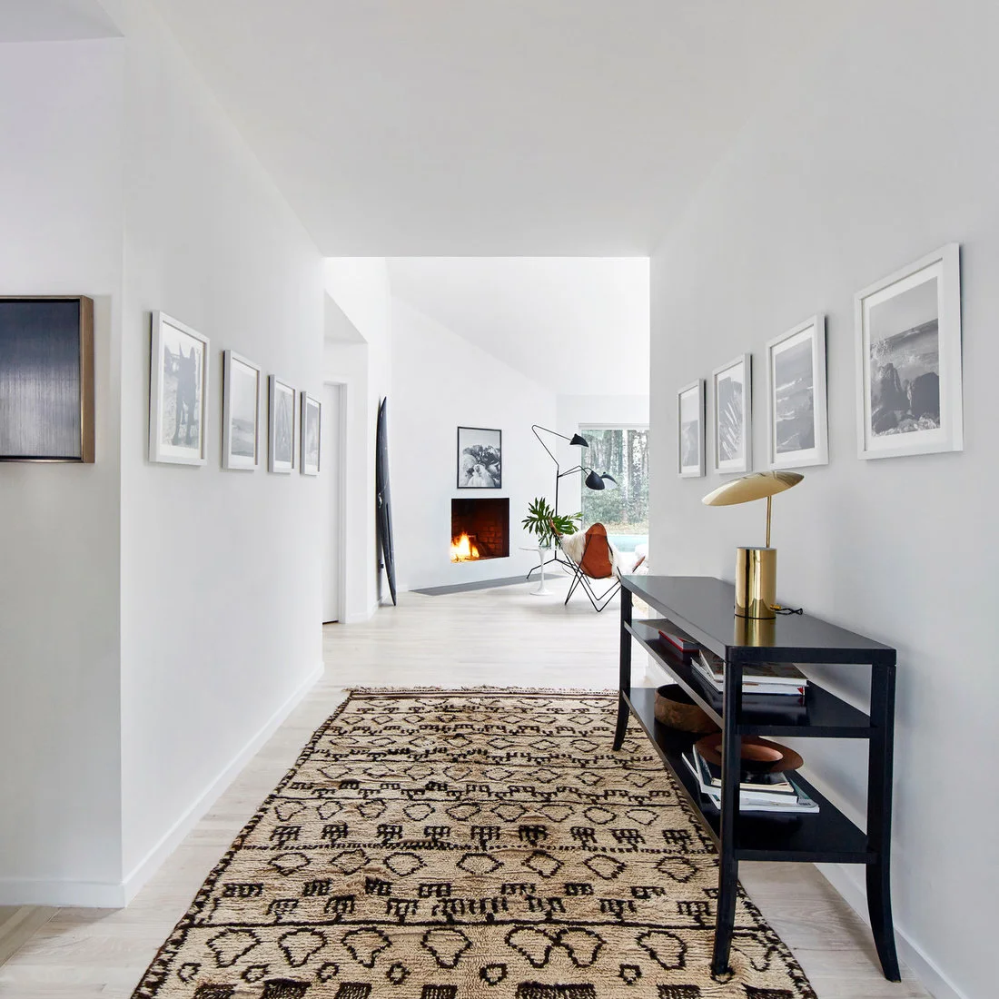 Rows of pale framed pictures line a white hallway above a dark console and patterned rug, with a living room visible beyond.
