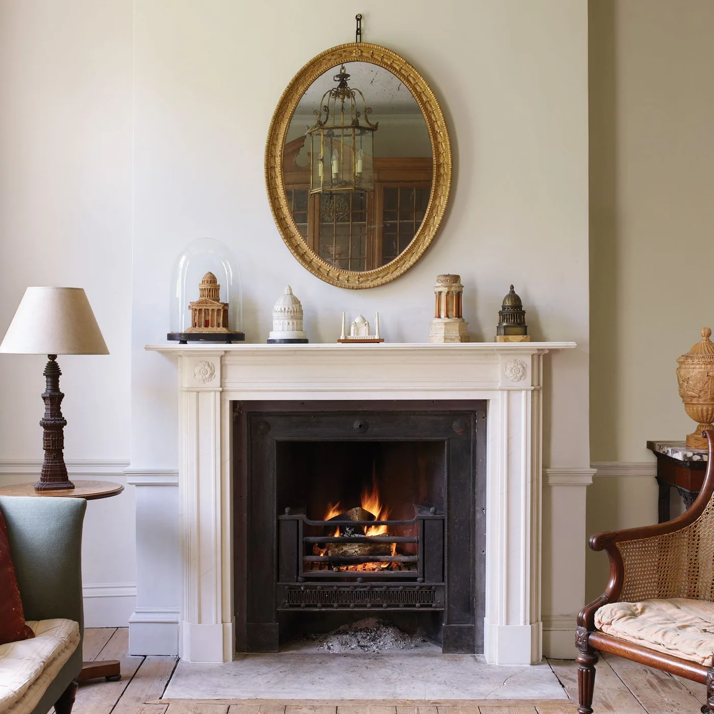 A pale fluted fireplace surround sits beneath an oval gold-framed mirror and a row of small architectural models.