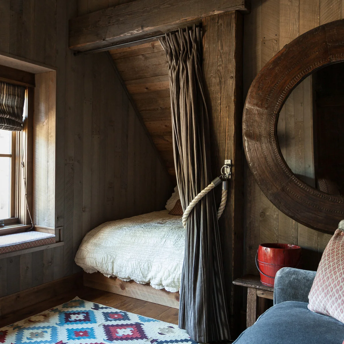 Wood-lined sleeping nook with pale bedding, a drawn-back curtain and colorful floor rug