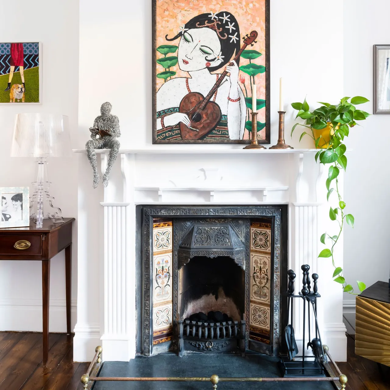 White mantel with two pale unlit tapers below colorful artwork beside a trailing plant.
