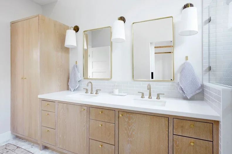 A pale wood double vanity stands beside a tall matching cupboard beneath two simple mirrors and three white wall lights on a pale tiled bathroom wall