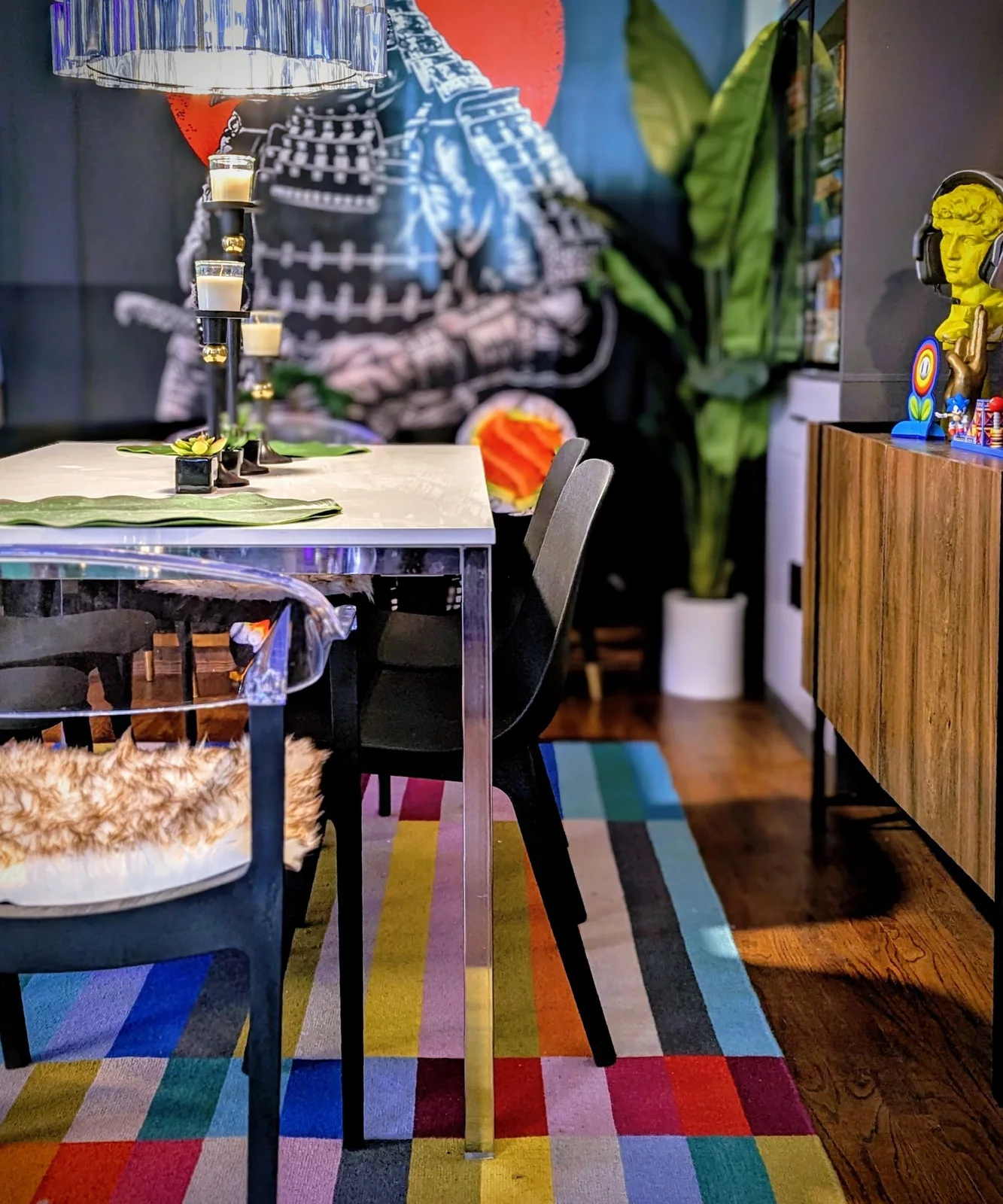 A reflective glass dining table stands beside slim dark chairs above a multicolored striped rug near a wood sideboard and patterned wall